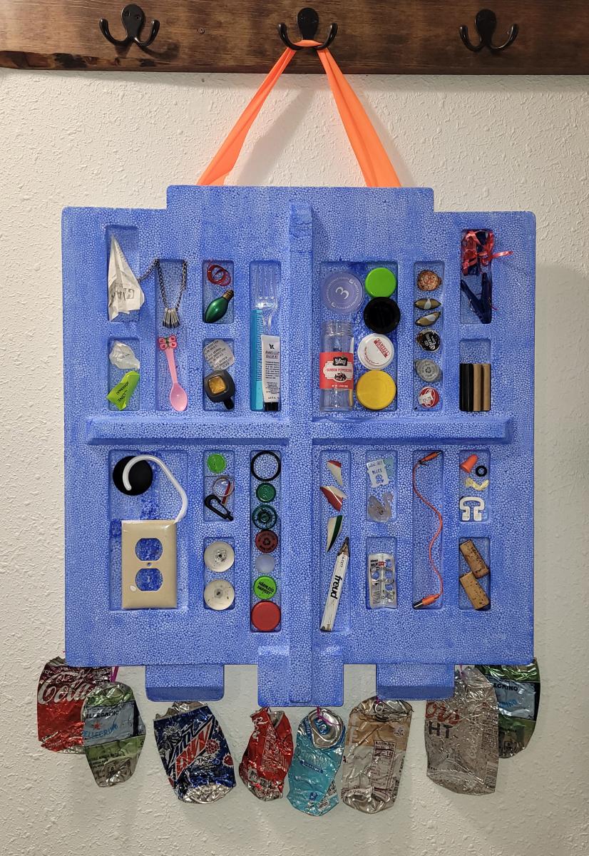 Blue packaging hanging from a hook with various litter items in the indents of the packaging. Crushed aluminum cans line the bottom.