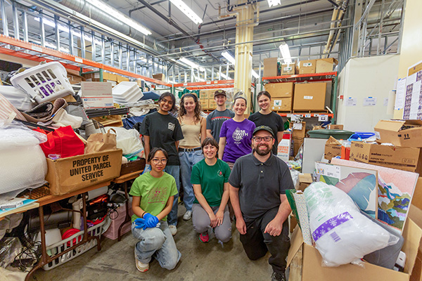 a group of people smiling in front of the camera in front of a warehouse with a lot of stuff