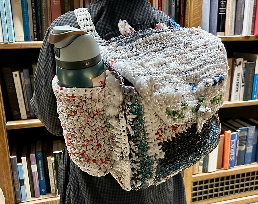 A handmade backpack woven from colorful plastic bags, with a thermos in the side pocket, worn by a person in a library