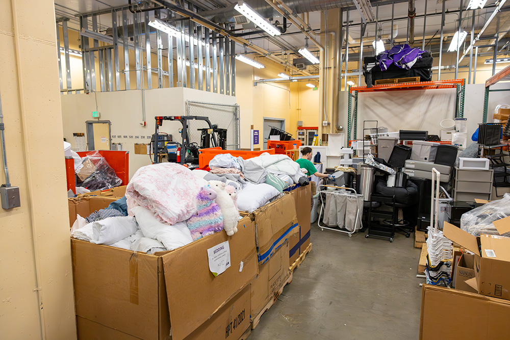 A small warehouse with several large cardboard boxes filled with bedding in the front. The corners of the warehouse have plastic storage containers lined up and other boxes.