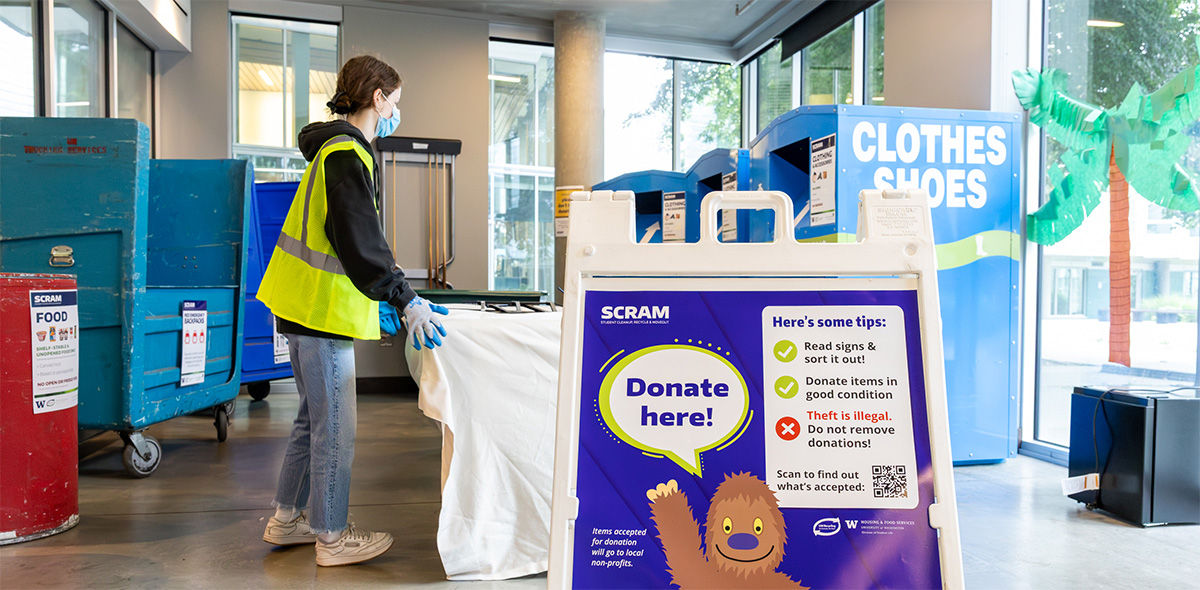 person in a high-visibility vest moving items on a table behind a sign that has an illustrated sasquatch that says "donate here!"