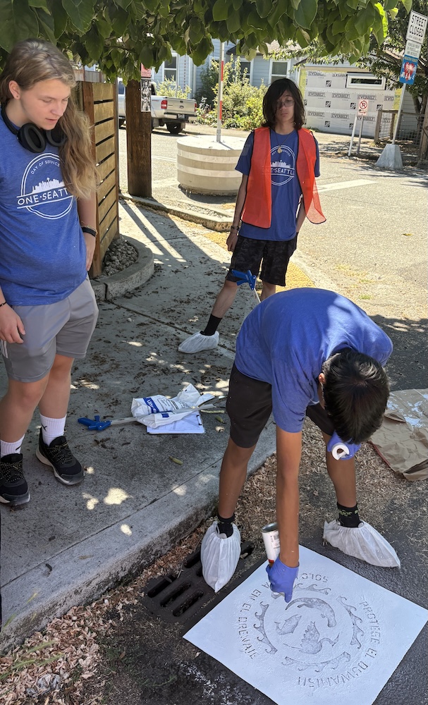 people painting a stencil by a stormdrain