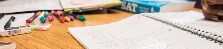 colored pens, erasers, post-it notes, notepads and a person's hand writing in an open notebook